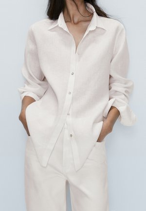 Kvinde iført løs hvid skjorte med knapper og op rullede ærmer samt hvide bukser, hænderne i lommerne, foran en ensfarvet baggrund.