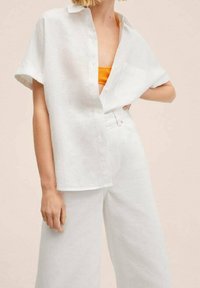 Chemise blanche en lin à manches courtes, boutonnée à l'avant, avec une poche sur la poitrine, associée à un pantalon blanc à jambes larges et un débardeur orange en dessous.