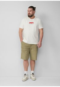 Witte katoenen T-shirt met "JAWS" grafiek in het rood, gecombineerd met kaki shorts. Witte sneakers met beige accenten en crew sokken. Casual fit.