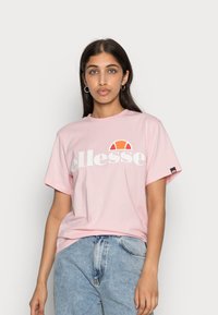 Light pink short-sleeve t-shirt with white "ellesse" logo and orange accent. Made from soft cotton, relaxed fit, with a tag on the sleeve.