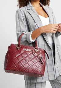 Sac à main bordeaux à motif avec texture brillante matelassée, logo argenté et détail pendentif ; doté de doubles poignées et d'une base plate pour plus de stabilité.