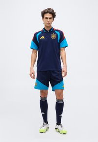 Jaunas vyras stovintis tiesiai, vilkintis tamsiai mėlyną Ispanijos futbolo aprangą su ilgomis kojinėmis ir futbolo bateliais, prieš balto fono foną.