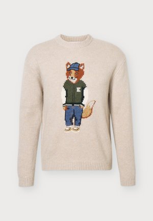 Un homme porte un pull crème orné d’un motif de renard, un jean bleu ample et des mocassins noirs sur un fond clair uni.