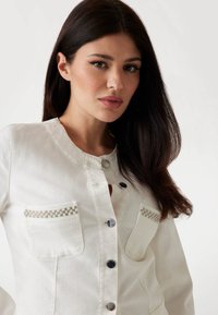 White denim jacket featuring silver button closure and pearl accents on pockets. Smooth texture with a fitted silhouette.