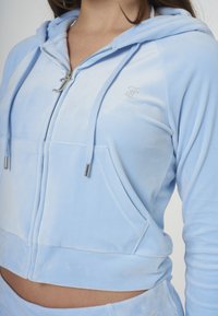 Casaco azul claro, com corte curto, feito de tecido macio. Apresenta fecho de correr frontal, dois bolsos, ferragens prateadas e um logótipo em strass no peito.