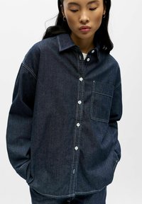 Donker denim overhemd met knopen, een kraag, een zak zonder klep en witte stikseldetails. Losse pasvorm met lange mouwen en knoopsluiting aan de voorkant.