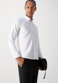 Camisa branca de manga longa com botões, feita de um tecido suave. Apresenta um colarinho clássico e detalhes de botões discretos. Combinada com calças pretas.