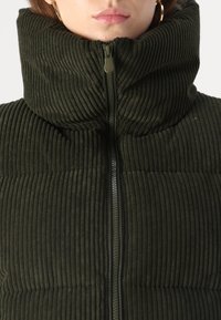 Vesta îmbrăcată, de culoare verde închis, cu guler înalt, dotată cu o textură striată și închidere prin fermoar frontal. Accentuata cu un fermoar solid.
