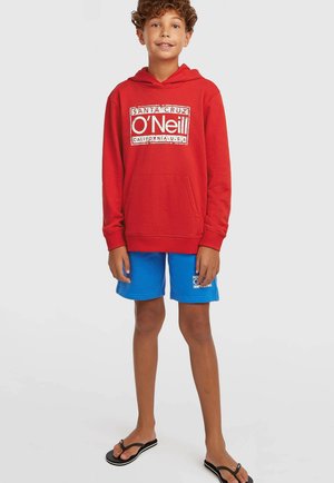 Czerwona bluza z kapturem z przednią kieszenią i białym logo. W komplecie niebieskie szorty z małą naszywką z logo. Noszone z czarnymi japonkami.