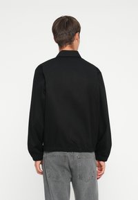 Svart jacka med krage, långa ärmar och elastisk nederkant. Slät textur, enkel design, bärs med grå jeans.