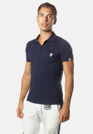 Man draagt een marineblauw poloshirt met leeuwenlogo en witte broek met trekkoord met "Leone"-branding, staand tegen een effen achtergrond.