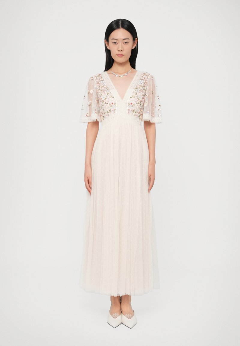 Μακρύ ελεφαντόδοντο φόρεμα με διάφανα κεντημένα μανίκια, λαιμόκοψη σε σχήμα V και υφασμένο μπούστο. Η αέρινη φούστα από τούλι έχει πουά μοτίβο.