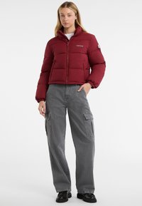 Donna che indossa un giubbotto invernale bordeaux, pantaloni cargo grigi e scarpe nere, in piedi con una mano in tasca su uno sfondo bianco.