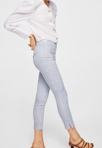 Bílá košile s knoflíky a balónovými rukávy, spojená s kalhotami v světle modré a bílé pruhy ke kotníkům, které mají boční rozparky, a hnědými sandály.