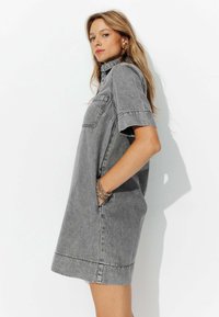Femme aux cheveux blonds portant une robe en denim grise à manches courtes avec une poche poitrine, se tenant les mains dans les poches, regardant sur le côté.