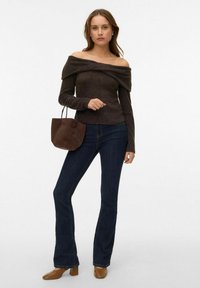 Pull en maille marron foncé à épaules dénudées, jean évasé bleu foncé, chaussures à talons marron, tenant un petit sac à main marron.