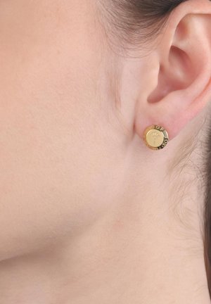 Linkes Ohr mit kleinem rundem goldenen Ohrstecker, graviert mit kreisförmigem Text und einem geflügelten Kaduzeus-Symbol in der Mitte.