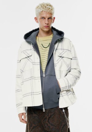 Bela karirana srajca z jopico v sivi barvi pod njo, plastena nad črtasto majico. Model nosi rjave hlače z vzorcem paisley.