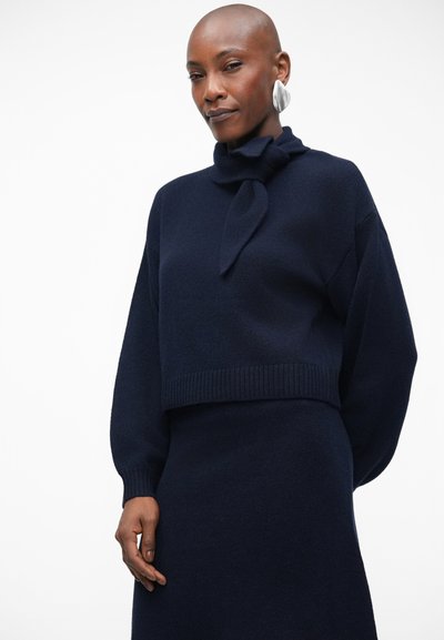 Sweater cropped azul-marinho com um detalhe de nó no pescoço, bainha canelada e mangas longas, feito de um tecido suave com uma textura lisa.
