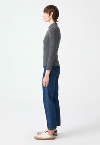 Profiilipildis naine, kes kannab halli soonikuga pikakäistega särki, siniseid teksaseid ja beeži-valgeid spordijalanõusid, seismas lihtsa valge tausta ees.