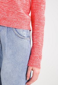 Maglione rosso lavorato a maglia con un modello testurizzato e polsini a coste, abbinato a jeans in denim azzurro chiaro con una tasca frontale.