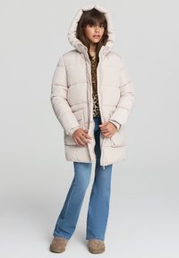 Puchowy, jasnoróżowy płaszcz zimowy z kapturem, z zamkiem błyskawicznym, prążkowanymi mankietami i bocznymi kieszeniami, noszony na bluzce w leopardzi wzór i niebieskich dżinsach.