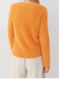 Vrouw in een donzige oranje lange mouwen trui, gecombineerd met lichtgekleurde broek, van achteren gezien tegen een effen achtergrond.