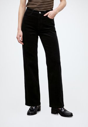 Persoon draagt zwarte ribfluwelen broek met hoge taille, een bruin-wit gestreept topje en zwarte loafers met hak, staat met één hand in de zak.