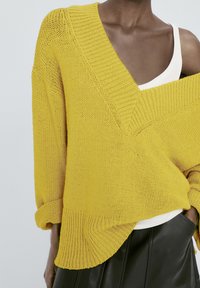Pull jaune tricoté avec un col en V plongeant et des manches retroussées, présentant un motif texturé et un ourlet incurvé. Porté par-dessus un haut blanc.