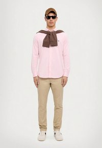 Ροζ πουκάμισο με κουμπιά, καφέ πουλόβερ περασμένο πάνω από τους ώμους, μπεζ παντελόνι, λευκά αθλητικά παπούτσια με μαύρες λεπτομέρειες, καφέ καπέλο και γυαλιά ηλίου.