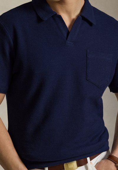 Άνδρας που φοράει μπλε μπλουζάκι πόλο με υφή και κοντά μανίκια, με τσέπη στο στήθος, λευκό παντελόνι και καφέ δερμάτινη ζώνη με χρυσή πόρπη.