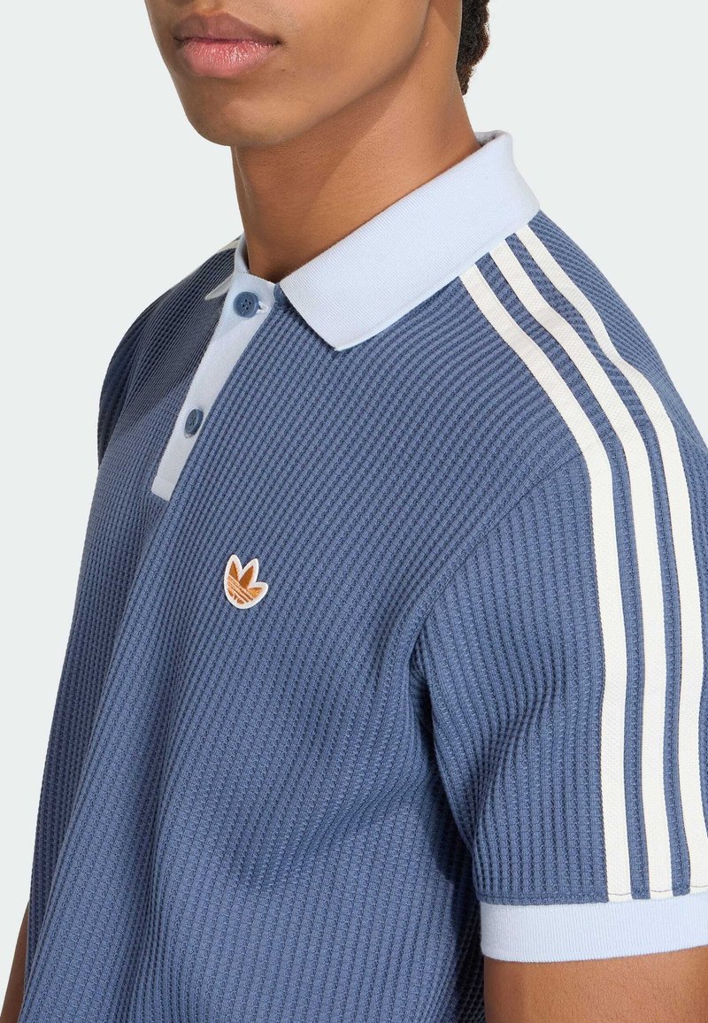 Uomo con polo a maniche corte blu con trama, colletto azzurro chiaro, strisce bianche sulle spalle e piccolo logo marrone sul petto.