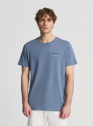 Giovane con capelli mossi che indossa una T-shirt blu a maniche corte con la scritta "WORK HARD", in piedi davanti a uno sfondo chiaro e uniforme.