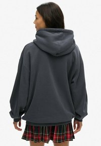 Sudadera gris de gran tamaño con capucha ajustable, tela texturizada y hombros caídos, combinada con una falda de cuadros rojos.