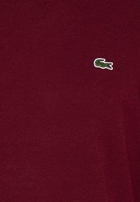Burgundy stickad polo skjorta med en texturerad tyg och en liten grön och vit broderad krokodil-logotyp på vänster sida.