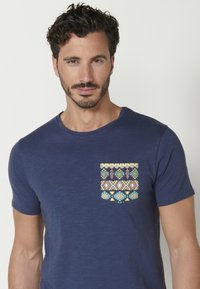 Námořnická modrá tričko vyrobené z bavlny s kontrastní kapsou s geometrickým vícetruncovým vzorem. Krátké rukávy, design s kulatým výstřihem.