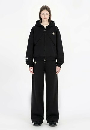 Joven mujer de pie de frente, vistiendo una sudadera negra con cremallera y emblema dorado, pantalones de pierna ancha a juego, zapatillas negras y fondo neutro.