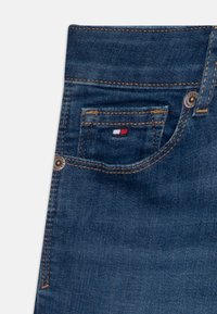 Jeans de mezclilla en un color azul oscuro, con un diseño clásico de cinco bolsillos, y un pequeño parche de logo en rojo, blanco y azul. Detalles de costura reforzada.