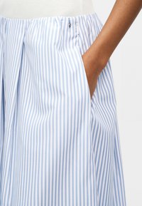 Χέρι με σκούρο χρώμα δέρματος στην τσέπη φούστας με κάθετες ρίγες σε ανοιχτό μπλε και λευκό, ψηλόμεσης, με πιέτες και πλευρικό φερμουάρ.