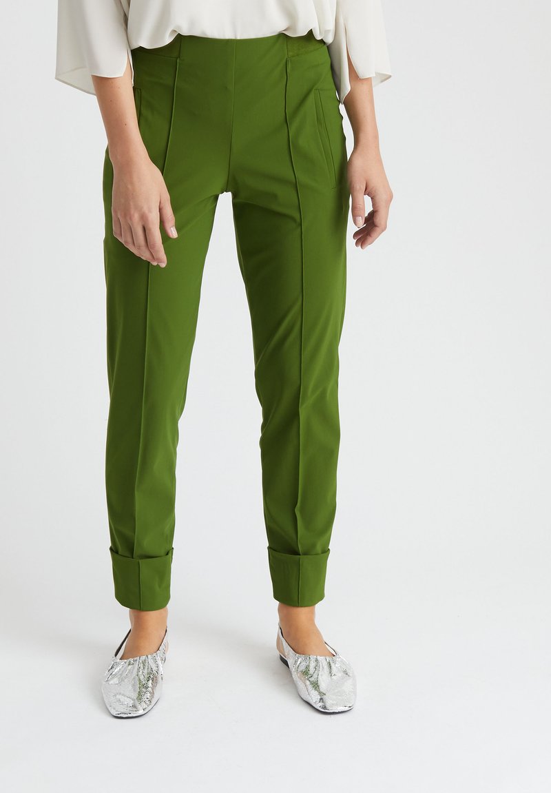 Grüne, maßgeschneiderte Hose mit hoher Taille, seitlichen Taschen und umgeschlagenen Säumen. Kombiniert mit silbernen flachen Schuhen. Glattes, leichtes Material.