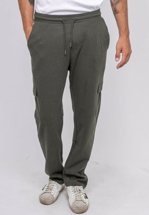 Homme portant un pantalon de jogging vert olive avec cordon à la taille, poches latérales et baskets blanches, debout sur un fond uni.
