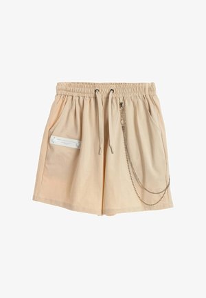 Pantalones cortos beige con cintura elástica y cordón, bolsillos laterales, detalle de etiqueta blanca en el bolsillo izquierdo y accesorio de cadena plateada adjunto en el lado derecho.