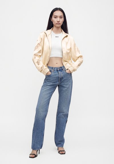 Jeune femme debout, mains dans les poches, portant une veste en satin beige, un top court blanc, un jean bleu et des talons ouverts noirs sur fond blanc.