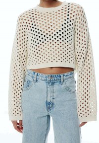 Pull court en maille ajourée crème avec manches longues, présentant un motif géométrique de trous et un accent noir sur la poitrine. Porté avec un jean bleu clair.