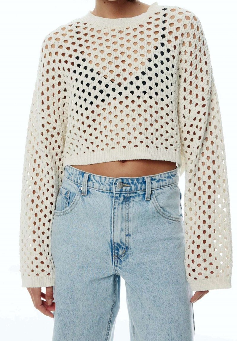 Pull court en maille ajourée crème avec manches longues, présentant un motif géométrique de trous et un accent noir sur la poitrine. Porté avec un jean bleu clair.