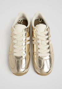 Sneakers in crème en goud met een glanzende metallic afwerking, vetersluiting en gestructureerde accenten. Voorzien van witte strepen aan de zijkanten.