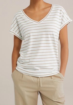 Vrouw die een wit T-shirt met V-hals draagt met dunne beige horizontale strepen en beige broek, met handen in de zakken.
