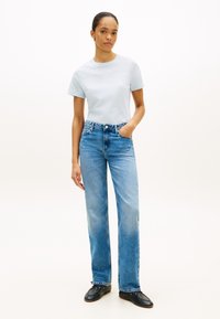 Ανοιχτό μπλε βαμβακερό μπλουζάκι με λογότυπο, συνδυασμένο με ψηλόμεσο μπλε τζιν. Το μοντέλο φοράει μαύρα μοκασίνια, στέκεται με το ένα χέρι στην τσέπη.