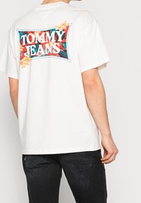 Camiseta blanca de algodón con un gráfico colorido de "TOMMY JEANS" con acentos florales en la parte posterior. Corte relajado y mangas cortas.