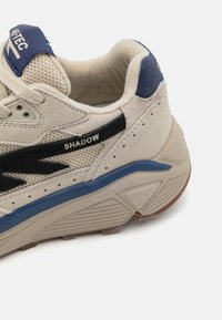 Adidași bej din materiale mesh și suede, cu accente negre, capac lateral al călcâiului albastru și talpă texturată. Include brandul pe limbă și pe lateral.